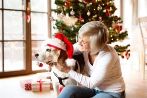 Beautiful senior woman sitting on the floor in front of Christmas tree with her dog wearing red Santa hat snuggling.